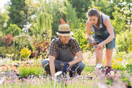 A gardener and a young girl are working together in a lush, well-maintained garden in Walthamstow. The gardener, wearing a straw hat, a checkered shirt, and grey trousers, is kneeling beside a bed of colorful flowers, which include pink, purple, and white blooms. The young girl, dressed in denim overalls and a grey t-shirt, is leaning forward, holding garden shears, and appears to be assisting with plant trimming. The garden features a neatly cut lawn in the foreground, bordered by flower beds with a variety of flowering plants and shrubs. In the background, there are mature trees, including a weeping willow, and several bushes, contributing to a green, leafy environment. The scene is set on a bright, sunny day with natural light illuminating the vibrant colors of the flowers and greenery, reflecting typical outdoor maintenance and gardening activities in Walthamstow. This image is relevant to landscaping and garden care services offered by Landscapers Walthamstow, highlighting professional gardening work in residential outdoor spaces.