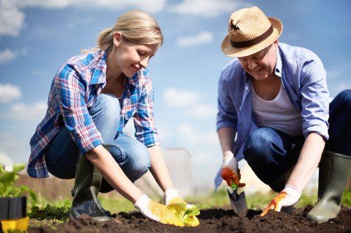 A young woman and an older woman are working together in a garden, planting or tending to a flower bed with dark, rich soil. The younger woman is wearing a blue plaid shirt and jeans, kneeling on the soil, holding a small plant or seedling. The older woman, dressed in a light blue shirt, dark jeans, and a wide-brimmed straw hat, is also kneeling and using a trowel to dig or position plants. They are both wearing gardening gloves and are focused on their gardening task, surrounded by a lush, green outdoor environment under a partly cloudy sky. The garden area appears well-maintained with visible edging and a natural setting suitable for gardening services in Walthamstow, with an emphasis on flower bed creation as part of landscaping or garden enhancement in the area.