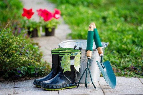 The image depicts a garden scene with gardening tools and accessories arranged on a paved pathway. In the foreground, there is a pair of black rubber gardening boots with a decorative pattern along the sole, positioned next to a pair of green-handled pruning shears and a small trowel with a blue handle, all resting on the paving. Behind these items, a white plastic bucket is partially visible, likely used for planting or watering. In the background, the garden features a lush, green lawn bordered by flower beds that include brightly colored pink and red flowers, and a few small potted plants are placed on the pathway leading into a well-maintained outdoor space. The scene is captured during daylight, with natural light illuminating the garden, which appears vibrant and lively, indicating it is the growing season or late spring. This setting suggests professional gardening or landscaping care by Landscapers Walthamstow, reflecting the natural elements of an active garden environment, and emphasizing outdoor maintenance and planting activities typical of gardening services in the Walthamstow area, near postcode E17.