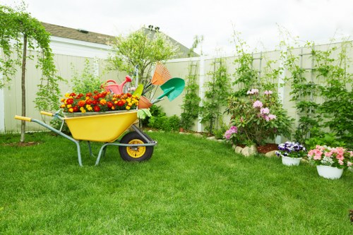 A well-maintained residential garden with a lush, green lawn bordered by flower beds containing colorful blooming plants and shrubs. In the foreground, a yellow wheelbarrow filled with gardening tools and decorative plastic flamingos sits on the grass. Towards the back, a white privacy fence encloses the outdoor space, with climbing plants and small trees growing along it. The garden features a variety of potted flowers in shades of pink, purple, and white, arranged along the edge of the flower beds. The landscape includes a mix of ornamental plants and natural foliage, creating a vibrant and inviting outdoor area suitable for gardening and landscaping services in Walthamstow. The scene is illuminated by natural daylight, suggesting a bright, partly cloudy day, perfect for outdoor maintenance or gardening work.