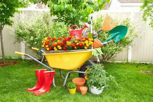 A brightly coloured garden scene featuring a yellow wheelbarrow filled with vibrant red, orange, and yellow chrysanthemums, positioned on a lush green lawn. Behind the wheelbarrow, there are various gardening tools and accessories, including a small green watering can, a red watering can, and a terracotta plant pot, alongside a potted plant with large green foliage. To the left, a pair of red rubber gardening boots rests on the grass. The background shows a white picket fence with dense shrubbery and trees, indicating a well-maintained outdoor space in Walthamstow, suitable for landscaping and garden maintenance services. The overall setting is bright and sunny, with natural light illuminating the scene, suggesting a pleasant spring or summer day in a typical UK suburban garden.
