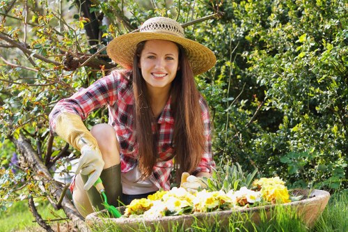 A young woman with long brown hair, wearing a straw hat, a red and white checked shirt, and gardening gloves, is kneeling in a lush outdoor garden during daylight hours. She is smiling while tending to a raised flower bed filled with blooming yellow and purple flowers, surrounded by green foliage and various garden plants. The background features dense bushes and trees with bright green leaves under natural sunlight, indicating a well-maintained outdoor space suitable for garden landscaping and lawn care services by Landscapers Walthamstow. The scene showcases a tidy garden layout with vibrant plant colors, natural textures of soil and greenery, and a peaceful environment, emphasizing outdoor maintenance activities typical of gardening services in Walthamstow postcode area.
