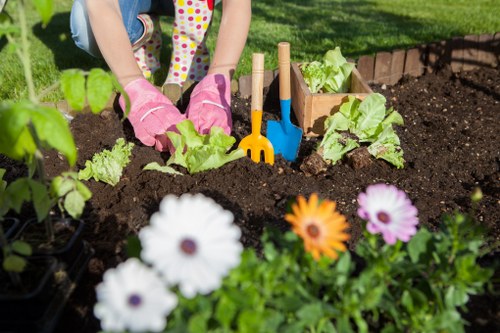 The image shows a person working in a well-maintained garden in Walthamstow, with gardening tools such as a small trowel and rake placed in the soil. They are wearing pink gardening gloves and are engaged in planting or tending to a vegetable bed that contains leafy greens, possibly lettuce or similar salad plants. In the foreground, there are colorful flower blooms, including white, orange, and purple flowers, adding vibrant natural tones to the scene. The garden features rich, dark soil with visible textures, bordered by a low brick edging, and a lush green lawn in the background. The environment appears sunny and well-kept, suggesting a typical outdoor gardening activity. This setting exemplifies basic gardening and planting services that a professional landscaper such as Landscapers Walthamstow might provide to create vibrant, healthy outdoor spaces in the local area around E17 postcode.