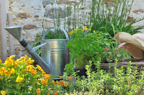 A small, vibrant garden scene featuring a variety of plants and flowers. In the foreground, there are bright yellow and orange nasturtiums with rounded leaves, contrasting with the lush green foliage of taller plants behind them. To the right, a potted plant with delicate pink flowers adds subtle colour variation. A rustic stone wall serves as the backdrop, partially visible behind the greenery. An aged, metal watering can with a long spout sits amidst the plants, suggesting regular garden maintenance. The garden area appears well-tended with a mixture of flowering plants, herbs, and foliage, creating a natural and inviting outdoor space suitable for gardening and landscaping activities in the Walthamstow area. The scene is illuminated by natural daylight, highlighting the textures and colours of the plants and garden tools, conveying a neat and productive small garden environment that complements professional gardening services offered by Landscapers Walthamstow.
