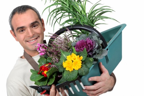 A smiling man with short dark hair and wearing a white shirt holds a green plastic garden fork filled with a variety of potted plants and flowers, including purple and yellow blossoms, in a landscaped outdoor garden setting. Behind him, a lush green plant with long, narrow leaves is visible, along with a section of grassy lawn and a paved patio area. The garden appears well-maintained with a mix of natural lawn, plant borders, and hard surfaces, suggesting professional gardening and landscaping care typical of services offered in Walthamstow, London. The overall scene showcases outdoor gardening activity suited for a residential backyard or front garden environment, under bright daylight with pleasant weather. This image is relevant for landscaping services provided by Landscapers Walthamstow and highlights the variety of plants and garden elements managed by local garden maintenance experts.