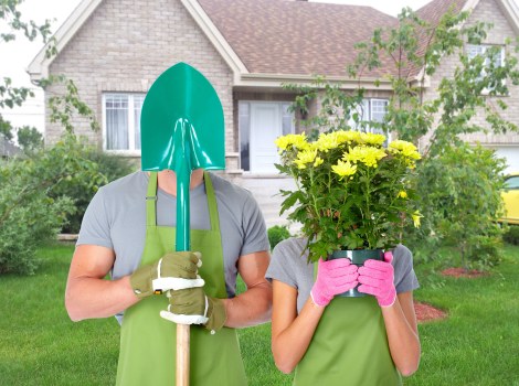 A garden scene featuring a well-maintained lawn with lush, green grass and a variety of shrubs lining the background. In the foreground, two individuals are standing on the grass, both wearing matching grey t-shirts, aprons, and gloves; one is holding a spade with a teal handle, and the other is holding a bright pink potted plant with yellow flowers. The garden area appears tidy with structured plant borders, and a brick house with white-framed windows and a front garden with small trees and flower beds is visible behind them. The weather looks clear, suggesting a pleasant outdoor gardening day. This scene reflects typical yard work and planting activities, relevant to professional gardening and landscaping services in Walthamstow, with natural lighting highlighting the vibrant greens and seasonal flowering plants, suitable for a landscaping company’s website focused on garden maintenance and planting projects.