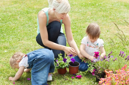 A woman with short blonde hair, dressed in a light blue sleeveless top and dark jeans, is kneeling on a well-maintained lawn in a garden, engaged in gardening activities with two young children. One child, wearing a white top and denim overalls, is bending down to examine or pick flowers, while the other, in a white dress with floral patterns, is sitting beside a flower bed, holding a small gardening tool. The garden features a lush green grass area, a flower bed with vibrant purple, pink, and orange blooms, and a few potted plants nearby. The background includes some trees and shrubbery, indicating a landscaped outdoor space typical of a domestic garden. The scene appears to be on a bright, clear day, providing natural daylight that highlights the fresh and lively environment. Landscaping and gardening services from Landscapers Walthamstow may assist in maintaining such vibrant and tidy outdoor spaces within the Walthamstow area, Suffix postcode E17, ensuring healthy plants and well-kept lawn and flower beds.
