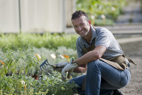 A smiling man wearing a grey shirt, brown apron, and gardening gloves is kneeling beside a garden bed in a backyard or landscaped outdoor space. He is holding a small garden rake and appears to be tending to green leafy plants and flowers, some of which are orange and yellow. The garden features a well-maintained lawn with dense grass in the foreground, bordered by soil and paving stones. In the background, there is a wooden fence and blurred greenery, suggesting a private outdoor area in Walthamstow. The lighting indicates a clear, sunny day, highlighting the natural colours of the plants and the tidy garden layout. This scene exemplifies outdoor maintenance and gardening activities typical of professional landscaping services offered by Landscapers Walthamstow, aligned with lawn care and garden upkeep in the local area.